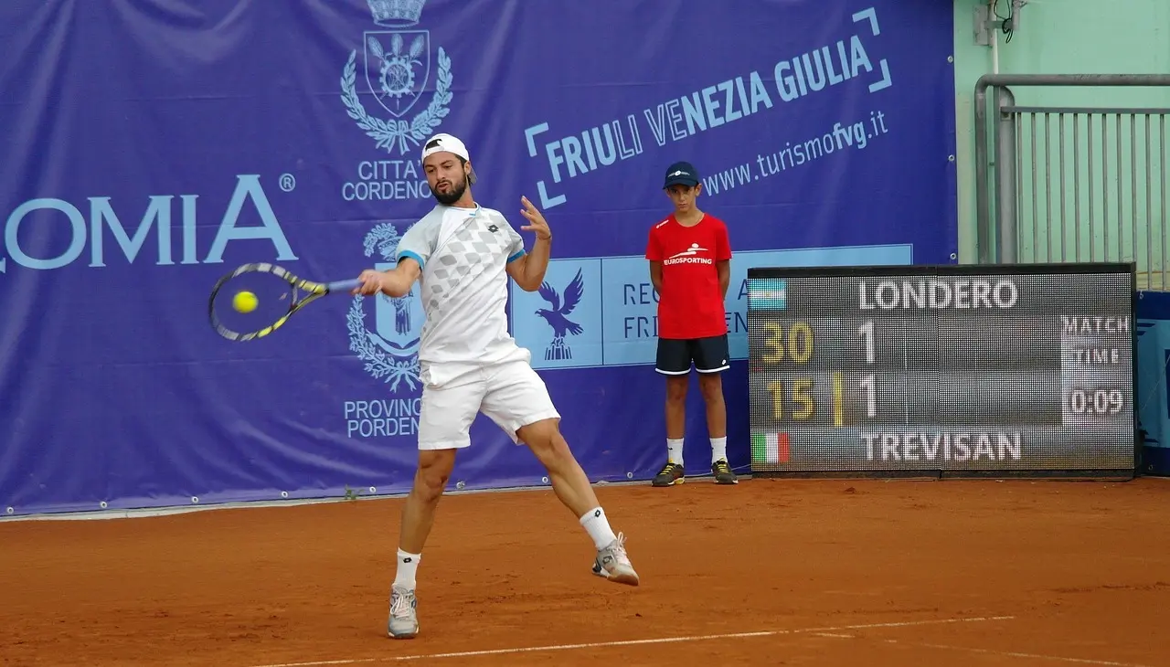Tennisspiller der slår en forehand på en grusbane med scoreboard i baggrunden
